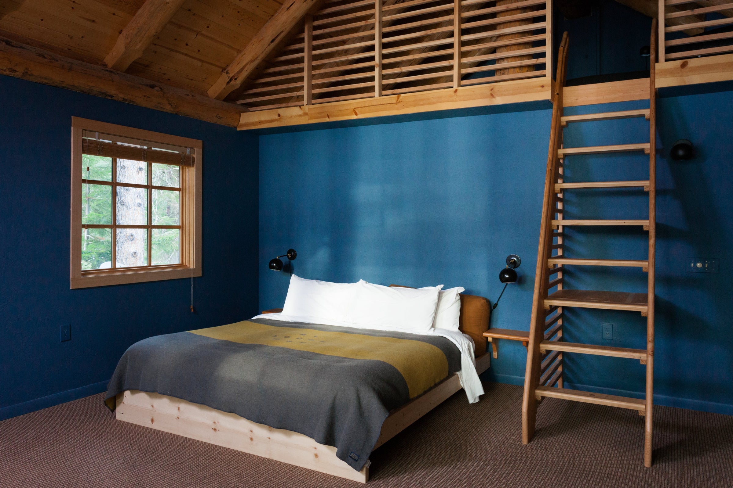 Cozy bedroom with low wood platform bed, white pillows, blue painted walls and a wooden ladder leading to a loft; window showing trees outside
