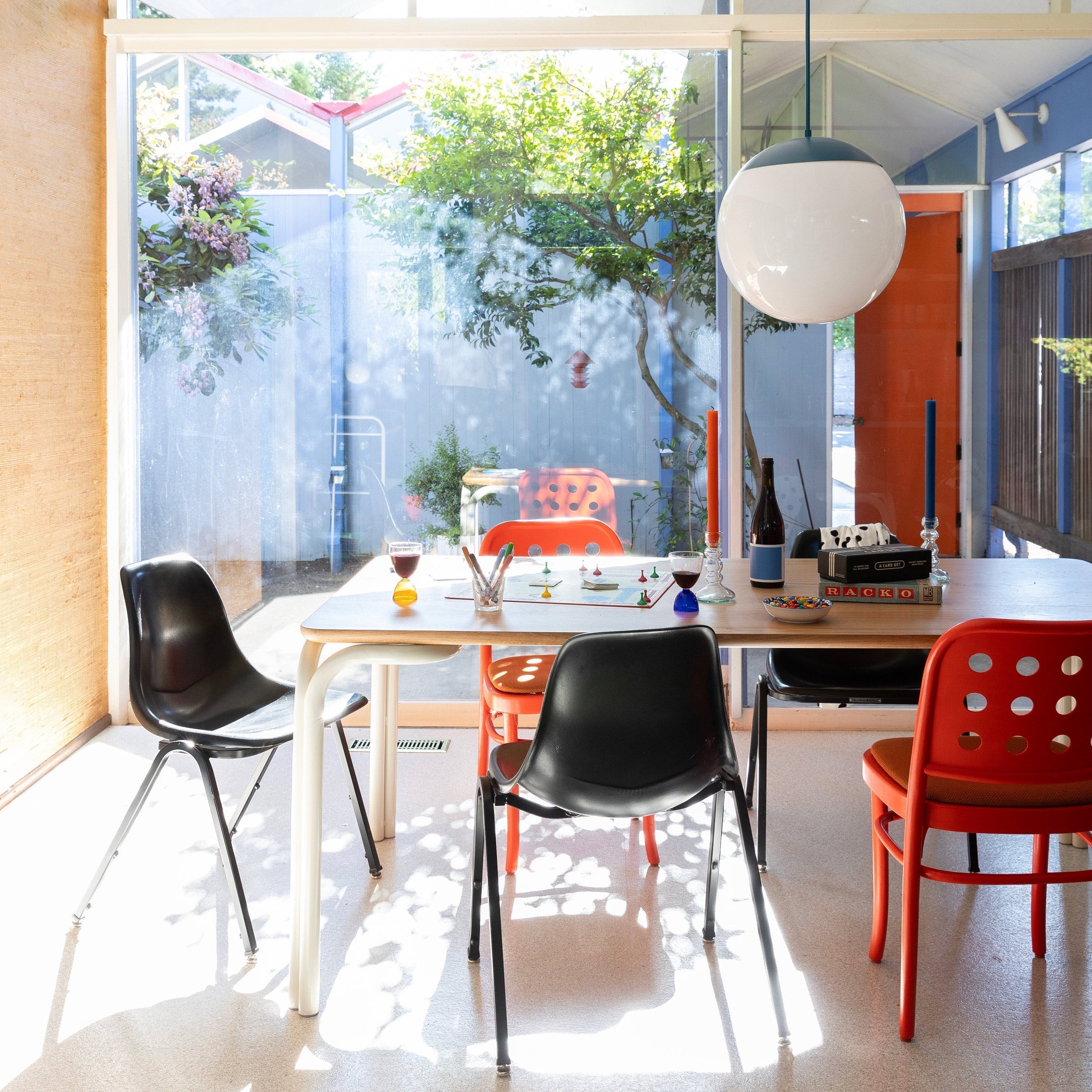 Sunlit dining table with mixed black and red chairs, a board game and wine glasses under a large round pendant light in front of a glass wall