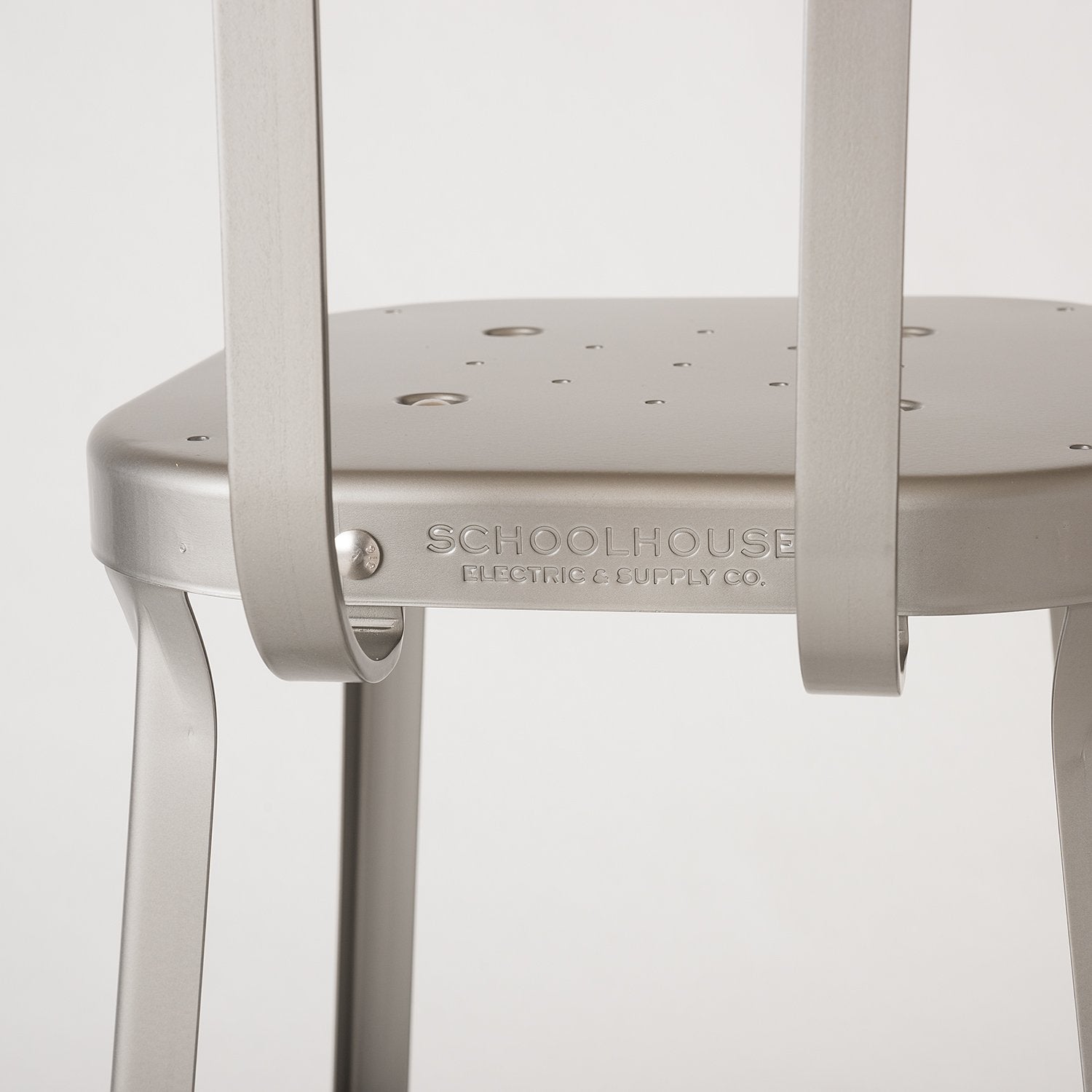 Close up of Utility Stool seat and back showcasing embossed SCHOOLHOUSE logo on raw coated steel with neutral background