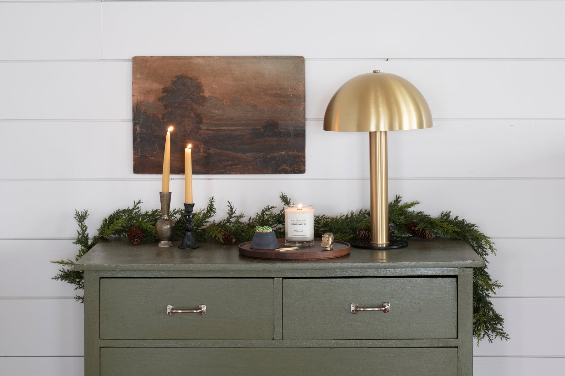 Dresser top with a lit Wildwood jar candle, two taper candles in vintage holders, and a brass dome lamp against white shiplap
