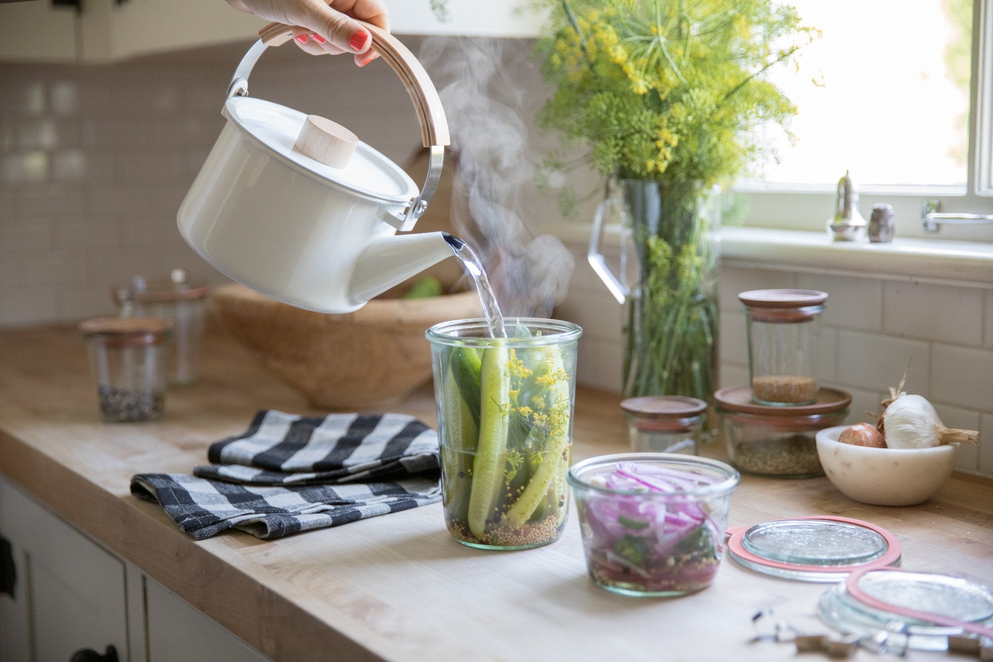 Hands pouring steaming water from a white kettle into a jar of cucumbers and dill for quick pickles on a sunlit kitchen counter