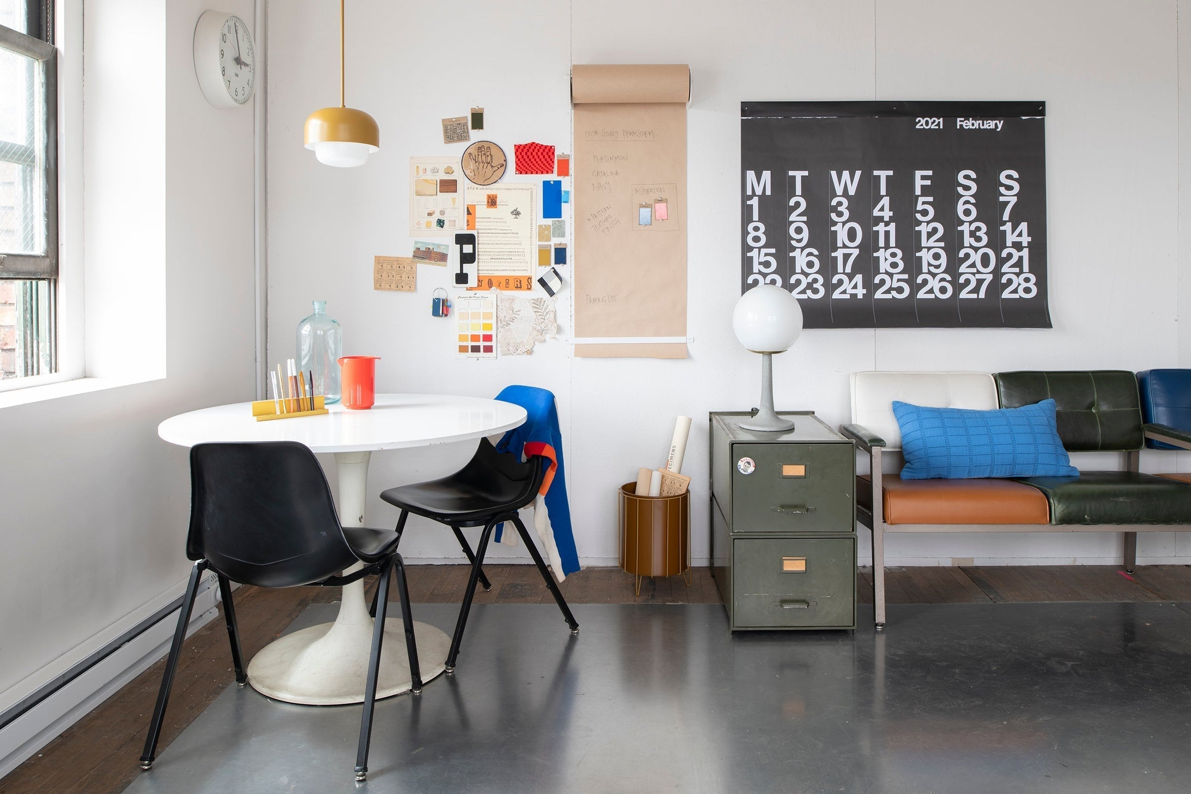 Sunlit studio seating area with round white table and black chairs, bulletin board, February 2021 wall calendar, metal filing cabinet and bench