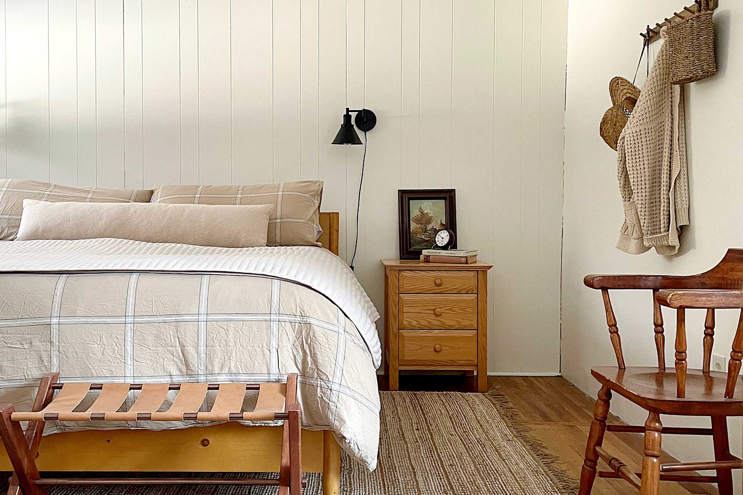 Cozy cabin bedroom with beige plaid bed, white paneled wall, wooden nightstand with framed art and alarm clock, woven rug
