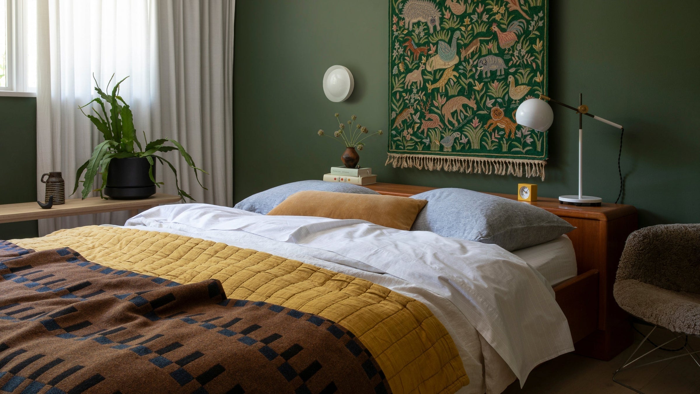 Bed with mustard quilt and patterned brown throw in cozy bedroom with green walls, textile wall hanging and bedside lamp