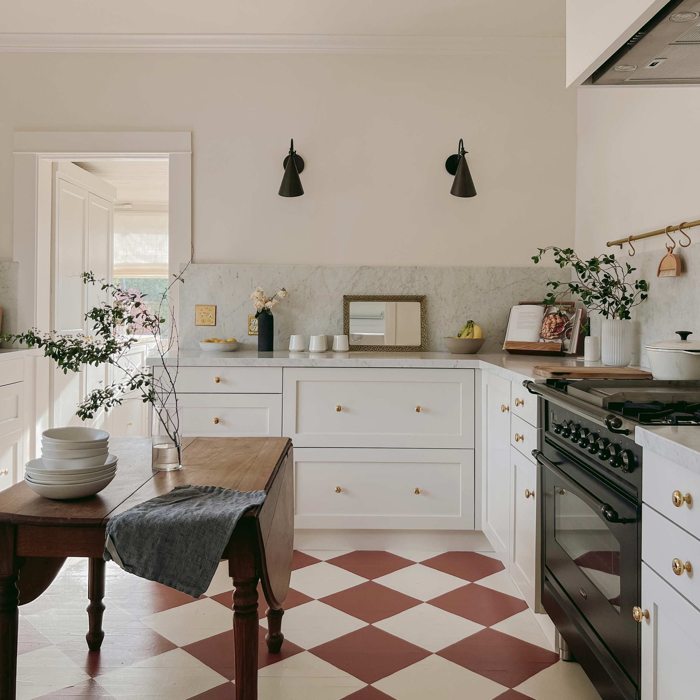 Sunlit kitchen with white shaker cabinets, marble backsplash, wooden island, black cone sconces and red checkerboard floor