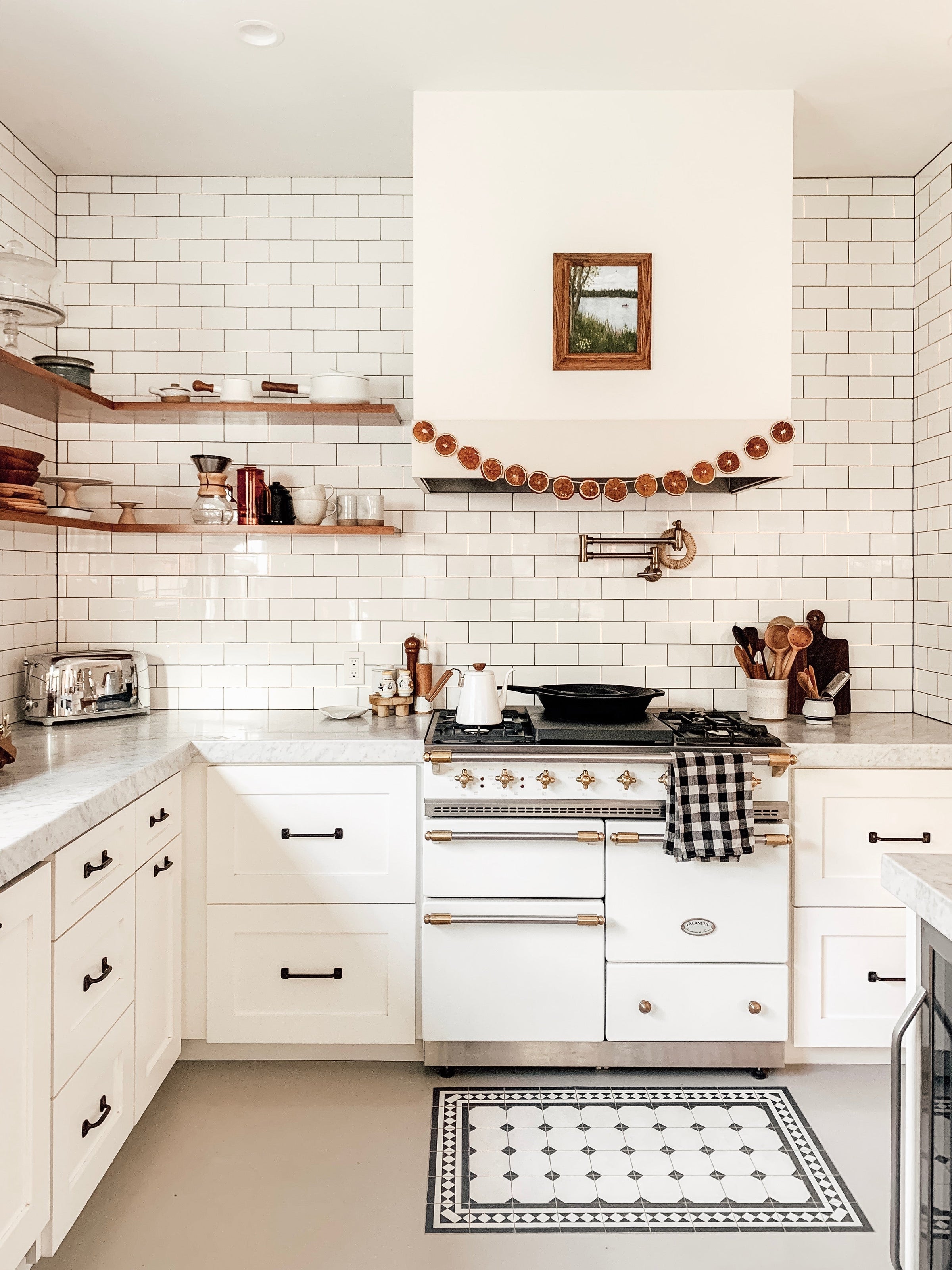 Bright farmhouse kitchen with white subway tile, open wood shelves, a white range with brass accents, and dried orange slice garland